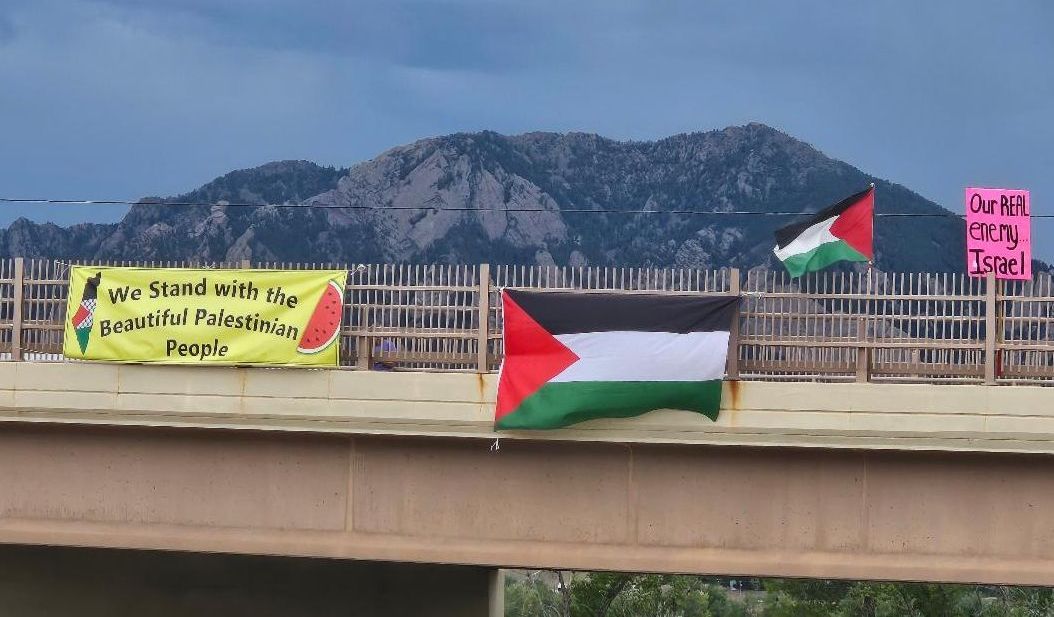 Two banners and Palestinian flags hung from the Cherryvale overpass, with the Flatirons behind.